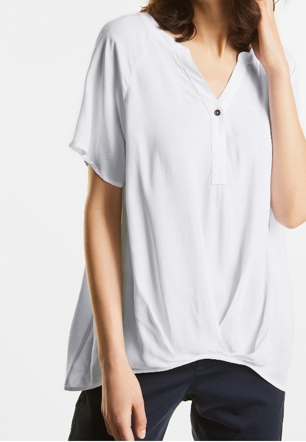 Femme portant un chemisier blanc ample à manches courtes avec un col en V et un bouton noir unique, ainsi qu'un pantalon bleu marine, la main touchant son cou.