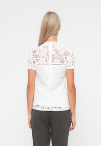 Donna bionda vista di spalle che indossa una blusa bianca in pizzo con maniche corte e pantaloni grigio scuro, su sfondo bianco.