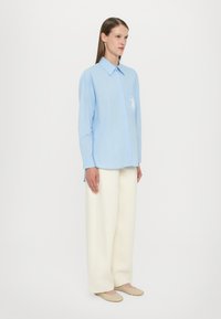 Vaaleansininen napitettu paita, jossa on pitkät hihat ja tasku, jossa on broderaita. Yhdistetty leveiden, vaalean cream-väristen housujen kanssa.
