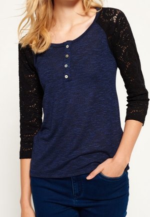 Vrouw draagt een blauw henley-shirt met zwarte kanten driekwartmouwen en donkerblauwe jeans, één hand in de zak.