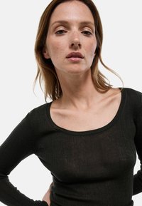 Haut à manches longues noir à côtes avec un grand décolleté arrondi, coupe ajustée et texture douce. Légère transparence dans le tissu visible.