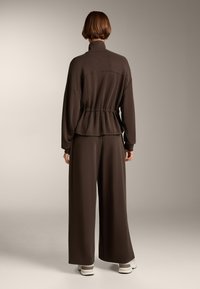 Tuta marrone con colletto alto e dettaglio in vita arricciata. Pantaloni a gamba larga, realizzati in un tessuto morbido, presentano una texture liscia.