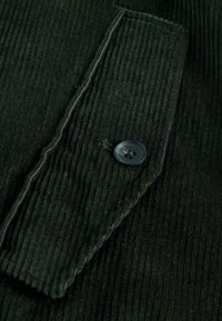 Donkergroene corduroy stof met een verticale ribbeltextuur, met een knoopsluiting bij de pocket en contrasterende stiksels langs de randen.