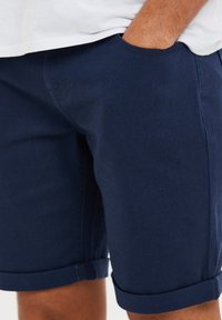 Marineblaue Baumwollshorts mit geradem Schnitt, rollierten Säumen und seitlichen Taschen, die einen glatten, strukturierten Stoff präsentieren.