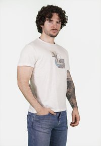 T-shirt in cotone color avorio con maniche corte, caratterizzata da una grafica dai colori beige, blu e nero sul fronte.