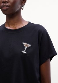 Sort bomulds T-shirt med et perlebroderet cocktailglasdesign i sølv- og guldtoner på venstre bryst. Korte ærmer og rund halsudskæring.