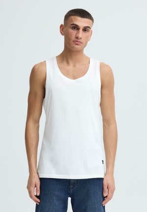 Jeune homme aux cheveux courts portant un débardeur blanc uni sans manches et un jean bleu foncé, debout devant un fond gris clair.