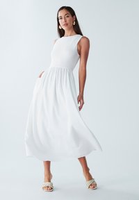 Robe maxi blanche avec un corsage texturé, une jupe fluide et lisse, et un design sans manches. Assortie à des sandales à brides tressées.