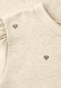 Tissu beige avec de petits cœurs gris foncé brodés près d'un col rond et un détail de manche froncée.