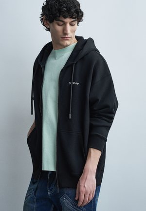 Ung mand med krøllet hår iført en sort zip-up hoodie over en lysegrøn t-shirt og blå jeans, kigger ned mod sin venstre side.