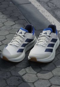 Chaussures de running Adidas blanches et bleues avec tige en mesh et semelles épaisses posées sur un pavé en pierre hexagonal près d’un bord de trottoir.