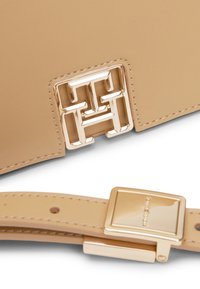 Borsa a tracolla in pelle beige con una chiusura a logo geometrico dorato. Presenta una tracolla testurizzata con una fibbia accentuata in oro. Superficie liscia con bordi cuciti.