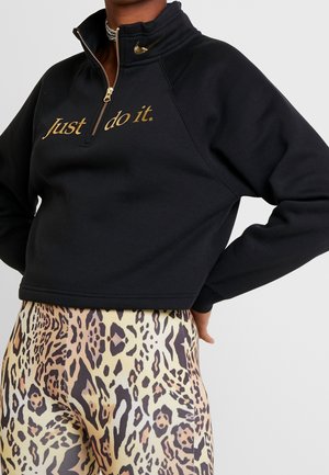 Czarna bluza z suwakiem do ćwiartki z złotym napisem i logo „Just do it.”, noszona z wysokim stanem legginsów w panterkę na osobie.