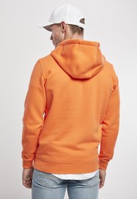 Sudadera naranja con un acabado texturizado, que cuenta con una gran capucha y puños acanalados, combinada con jeans de mezclilla azul claro. Gorro blanco en el fondo.