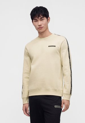 Jeune homme aux cheveux noirs courts portant un sweat beige et un pantalon noir avec des bandes de marque sur les manches et des écussons logo.