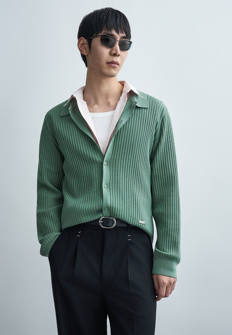 Πράσινη ριμπ ζακέτα με κουμπιά μπρ οστά, φορεμένη πάνω από λευκό μπλουζάκι με στρογγυλή λαιμόκοψη και ροζ πουκάμισο με γιακά, συνδυασμένη με σκούρο πλισέ παντελόνι.