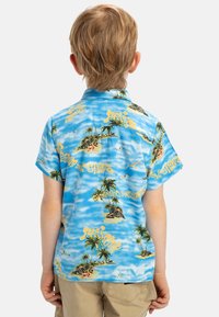Korte mouwen, lichtblauwe shirt met tropische eilandpatronen, gele tekst en afbeeldingen van palmbomen, gemaakt van een zachte stof.