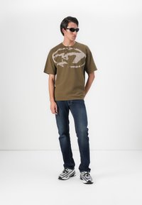 Olivově zelené tričko s krátkým rukávem a světlou grafikou, v kombinaci s tmavě modrými džíny a stříbrnými sportovními teniskami. Černé sluneční brýle na sobě.