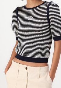 Pruhovaný pletený svetr v námořnické modři a bílé barvě s krátkými, vypouklými rukávy a zkráceným lemem. Na hrudi se nachází kruhové logo.