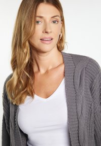 Grauer gestrickter Cardigan mit strukturierten Zopfmuster, kombiniert mit einem weißen V-Ausschnitt-Top. Das Design bietet eine lässige Passform und einen offenen Vorderteil.