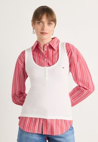 Hvit tanktop med ribbet tekstur, dyp utringning og knappe detaljer, lagt over en rød og hvit stripete dresskjorte med oppbrettede ermer.