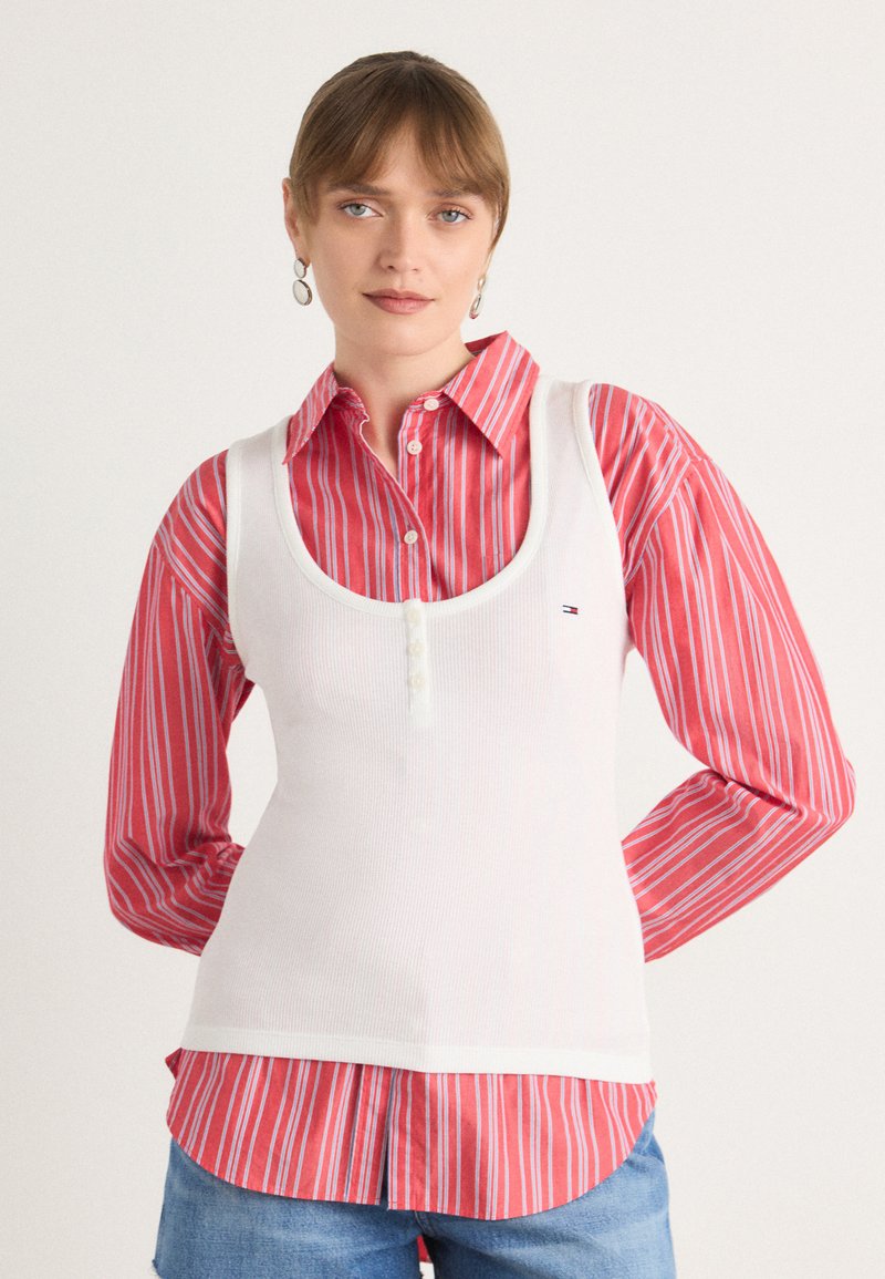 Hvit tanktop med ribbet tekstur, dyp utringning og knappe detaljer, lagt over en rød og hvit stripete dresskjorte med oppbrettede ermer.