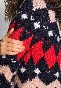 Pull en tricot avec un motif en zigzag en bleu marine, rouge, rose et blanc. Le tissu semble épais et texturé, avec une main reposant sur l'épaule.