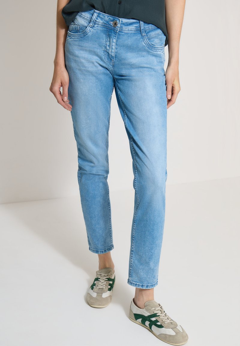 Femme portant un jean bleu clair et des baskets beiges avec des rayures vertes, debout, les mains détendues le long du corps sur un fond uni.