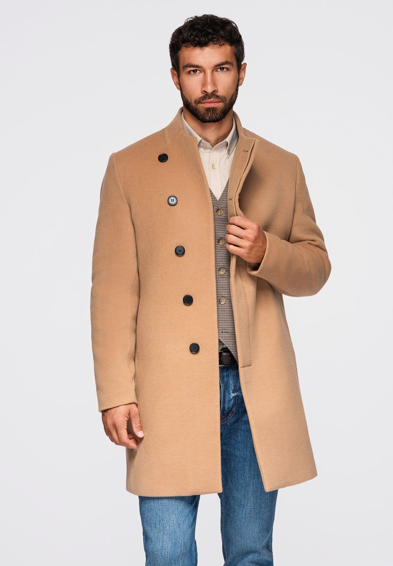 Mand iført en beige jakke over en beige skjorte og vest med blå jeans, stående mod en ensfarvet lys baggrund.