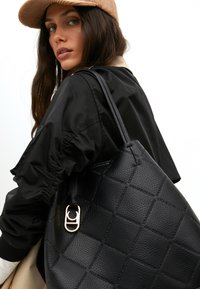 Eine Frau mit langen dunklen Haaren trägt eine braune Mütze und eine schwarze Jacke. Sie hält eine strukturierte schwarze Handtasche mit goldenen Tragegurten.