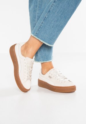 Baskets blanches avec des semelles en caoutchouc couleur caramel, matériau texturé et lacets plats. Présente un logo discret sur le côté.