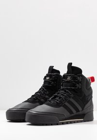 Baskets montantes noires en cuir et en daim, présentant une surface texturée, des semelles épaisses, et une boucle de talon rouge avec trois rayures noires.