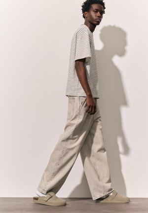 Jeune homme marchant, portant une chemise à manches courtes rayée, un pantalon beige ample, des chaussettes blanches et des chaussures beige à enfiler sur un fond uni.