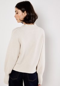 Sweter w kolorze kremowym z wysokim, prążkowanym kołnierzem, teksturowanym wzorem dzianiny i rękawami typu balloon, w połączeniu z ciemnoniebieskimi dżinsami.