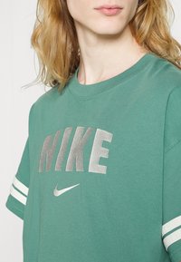 Grön bomullströja med silverfärgad "NIKE"-logotyp, rund halsringning och vita randiga detaljer på ärmarna. Slät textur och avslappnad passform.
