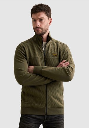 Homme aux cheveux bruns courts et à la barbe, portant une veste matelassée zippée vert olive, bras croisés, regardant légèrement sur le côté.