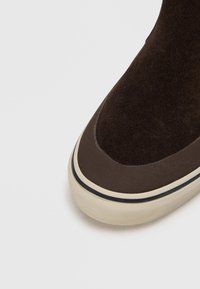 Brune suède høye sko med en mørkebrune gummisåle og kremfargede detaljer langs kanten. Har et slankt, sømløst design.
