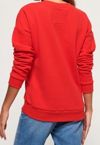Sudadera roja de manga larga, con puños acanalados y cuello redondeado. Presenta un pequeño parche en la parte superior de la espalda y un corte relajado.