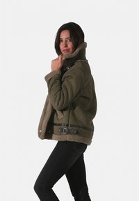 Veste en shearling vert olive avec un col haut, doublée de polaire beige, accents en cuir noir, et fermeture à boutons-pression. Jean noir porté.