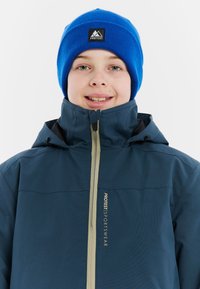 Lachend kind met een blauwe gebreide muts met logo en een dichtgeritste marineblauwe Protest sportjack met capuchon.