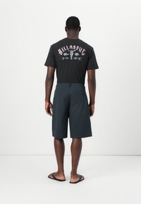 Zwarte t-shirt met roze grafische tekst en bloemdesigns op de achterkant, gecombineerd met donkere shorts en slippers. Eenvoudige en casual look.