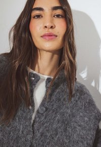 Cardigan gris, de textura esponjosa con botones redondos, superpuesto a un suéter blanco de canalé. El fondo es claro, enfatizando la prenda.