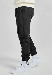 Person i svarta cargobyxor med ankelknyt, vita strumpor och vita Nike-sneakers, stående mot en enkel ljus bakgrund.