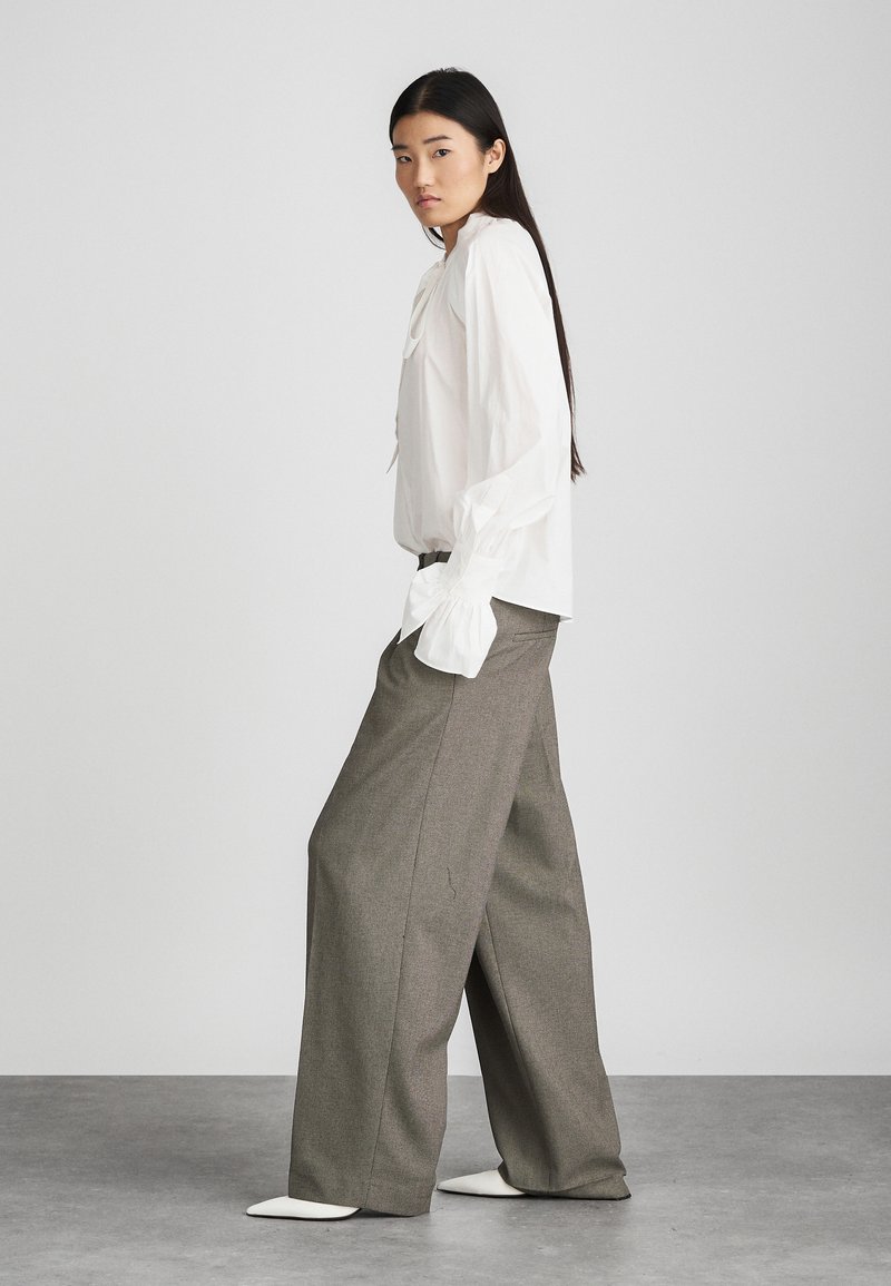 Blusa bianca con maniche a sbuffo e dettaglio con fiocco, abbinata a pantaloni grigi a vita alta e gamba larga, e scarpe bianche a punta.