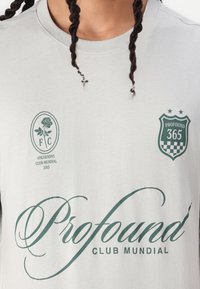 Lys grå t-shirt med mørkegrønne grafik, herunder "Profound" skrift, et rose emblem og et skjold med teksten "PROFOUND 365".