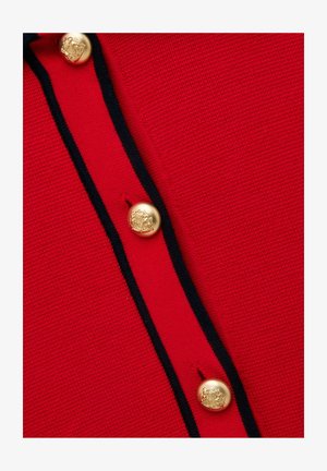 Detailný záber na červenú pletenú látku s gombíkmi lemovanými čiernou páskou, ktoré majú tri zlaté gombíky s reliéfnym motívom erbu.