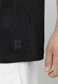 Tissu noir texturé d'une chemise, avec un design en relief et un logo subtil près de l'ourlet. Gros plan sur la partie inférieure de la chemise et la main.