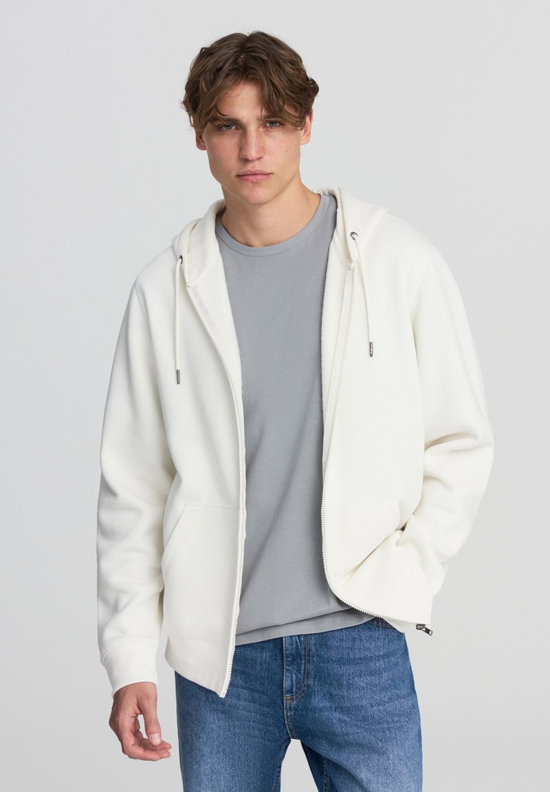 Felpa bianca con zip e coulisse, caratterizzata da una texture morbida. Indossata sopra una t-shirt grigia, abbinata a jeans blu. Il modello posa in modo neutro.