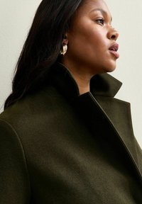 Cappotto in lana verde oliva con colletto alto e design strutturato; presenta linee pulite e una consistenza morbida. Orecchini a cerchio dorati aggiungono un tocco di dettaglio.
