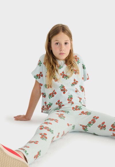 Jeune fille aux longs cheveux portant une tenue bleu clair avec des imprimés de cerises rouges, assise par terre avec les jambes étendues et les mains derrière elle.